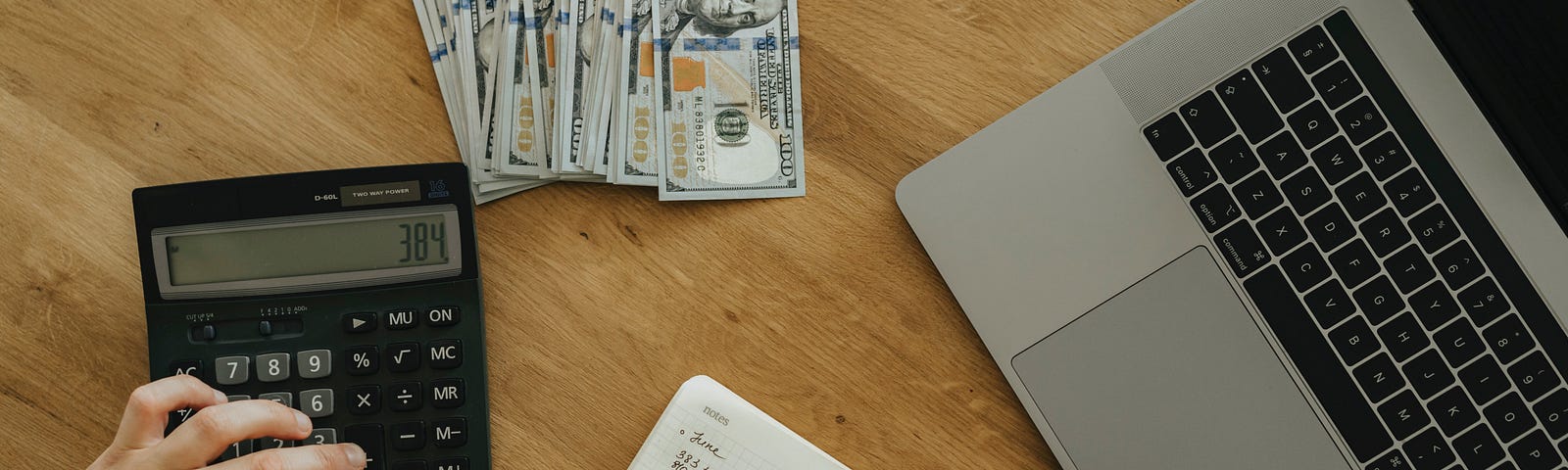 Here we have a picture of a person calculating all of the money that they made writing, just like I did for this article. There are disembodied hands over a calculator and notepad. A stack of money and laptop is nearby on a wooden table.