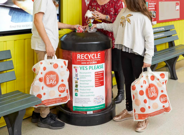A colourful photo of two children and an adult standing around a Coles REDcycle bin, shot so they can only be seen from the shoulders down to their feet. They are all placing rubbish in the hole at the top of the bin lid, which is rounded with the words ‘soft plastics’ embossed on it. The children each carry a reusable plastic Coles bag.