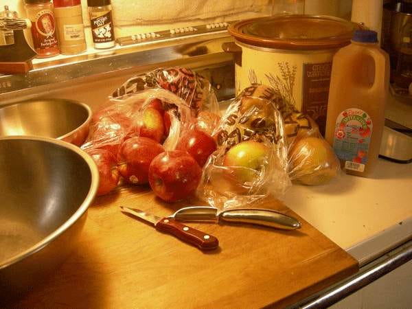 Two bags of apples, a half gallon container of apple cider, a paring knife, a vegetable peeler, two metal bowls and a slow cooker on a white stove topped with a wooden cutting board.