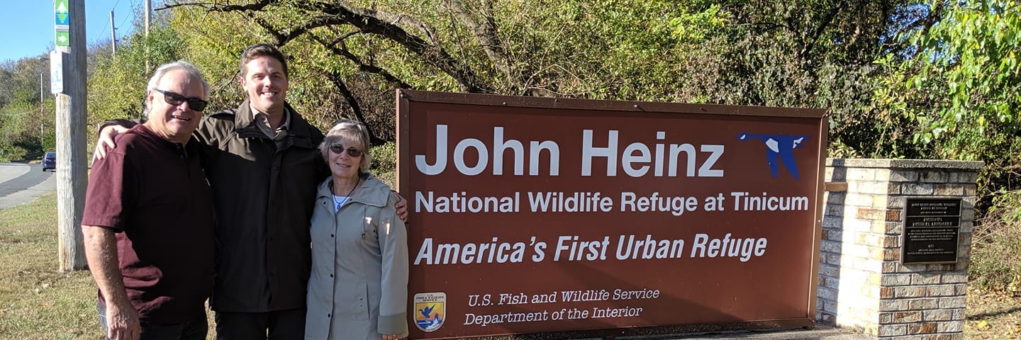 Kevin, David, and Cheryll stand together to the left of the John Heinz NWR welcome sign on a bright day.