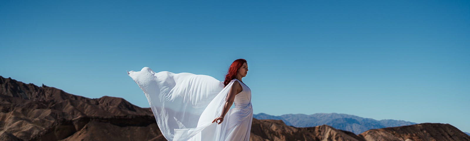 A woman dressed in a white flowing dress, which is blowing in the wind. She is standing on a rocky hill in desert terrain.