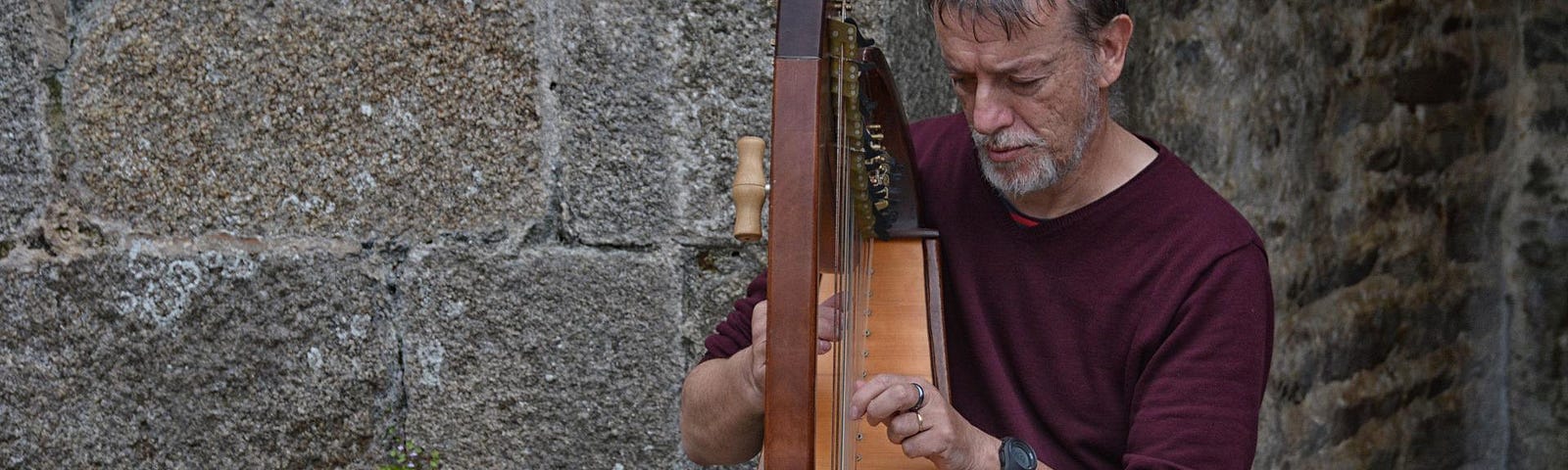 A man sits in front of a stone wall, playing a harp. He is wearing a maroon shirt and grey pants. He has a grey beard.