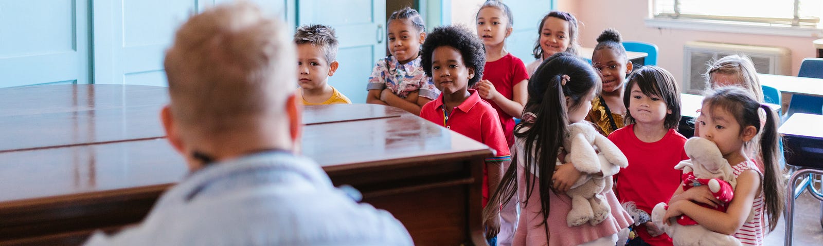 Children in a music classroom. They are standing in front of their desks and a male teacher is sitting at a grand piano.