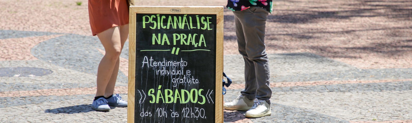 Na praça, de manhã, duas pessoas conversam entre uma placa que anuncia “Psicanálise na Praça: atendimento individual e gratuito aos sábados das 10h às 12h20”. O texto foi escrito com giz, em um quadro negro