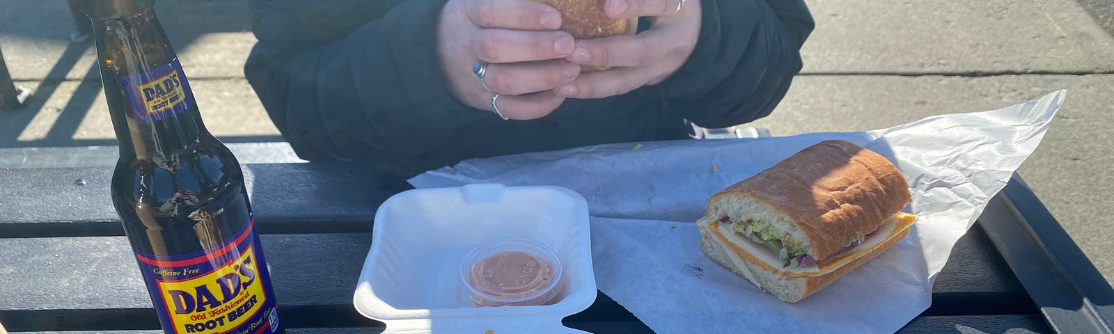 A person sits at an outdoor table holding half of a sandwich. The other half of the sandwich, some fries and a Dad’s Root Beer are on the table in front of them.