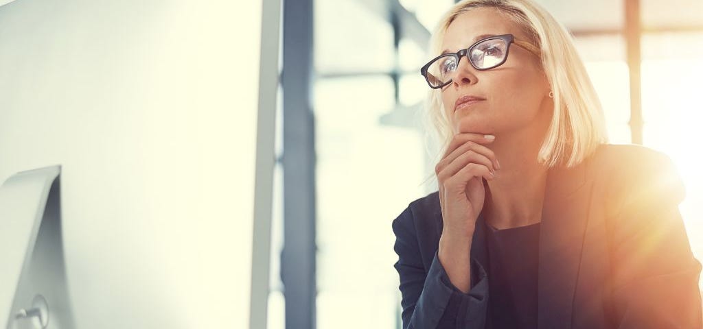 A blonde business woman in a dark suit sits facing a large computer screen.