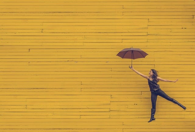 Yellow background, with a happy woman leaping and holding an umbrella as if it is causing her to fly.