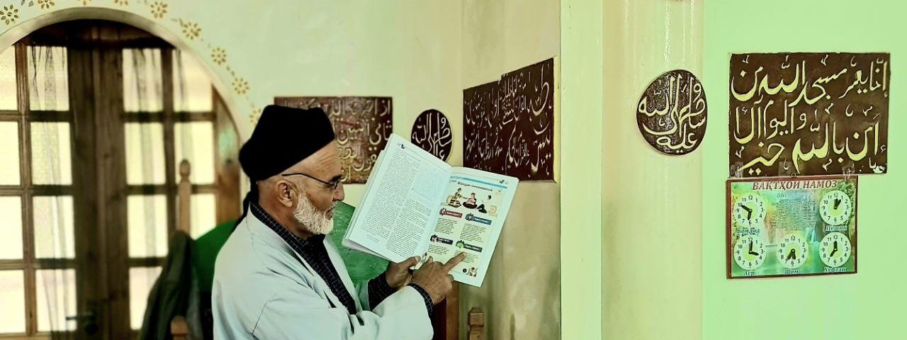Domullo Shermuhamad stands at the front of the mosque reading from a Healthy Mother, Healthy Baby booklet.