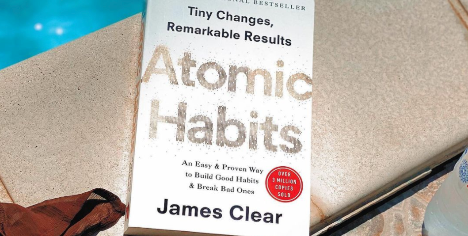 An image of the book ‘Atomic Habits’ by James Clear. The book is shown by the pool on holiday with great sunny weather!