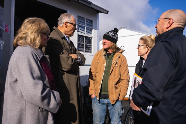Gov. Jay Inslee and Trudi are standing outside bundled up in warm jackets, chatting with a man in a stocking cap at a Longview tiny home village. The sun is shining while they talk.