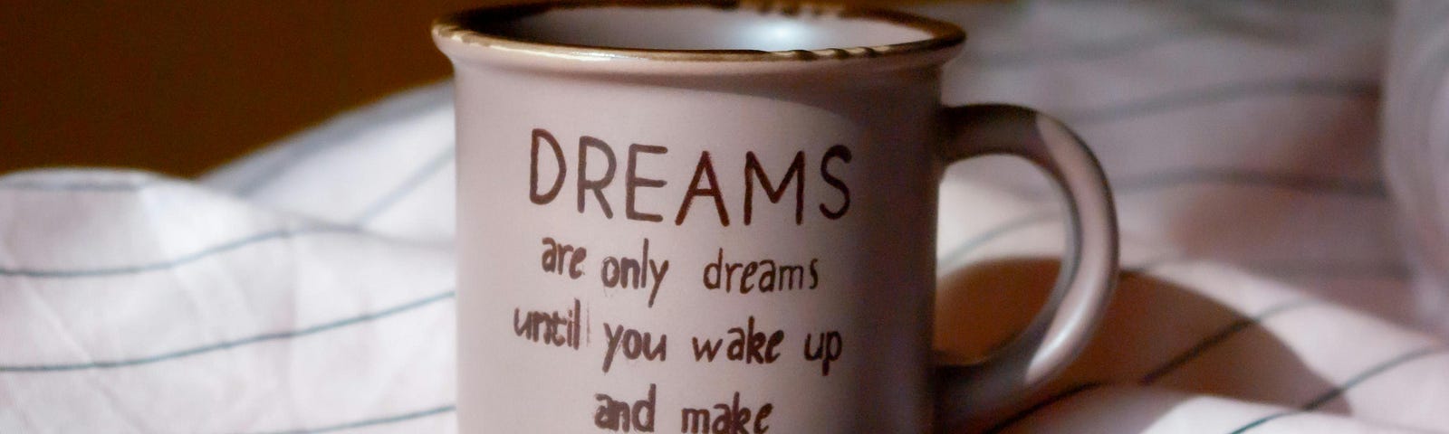 A white coffee cup sits on striped sheets and has a quote that reads, “Dreams are only dreams until you wake up and make them real”.