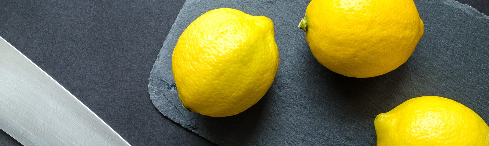 Three lemons on a countertop next to a large knife