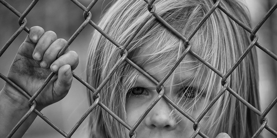 sad child looking out from behind wire fence