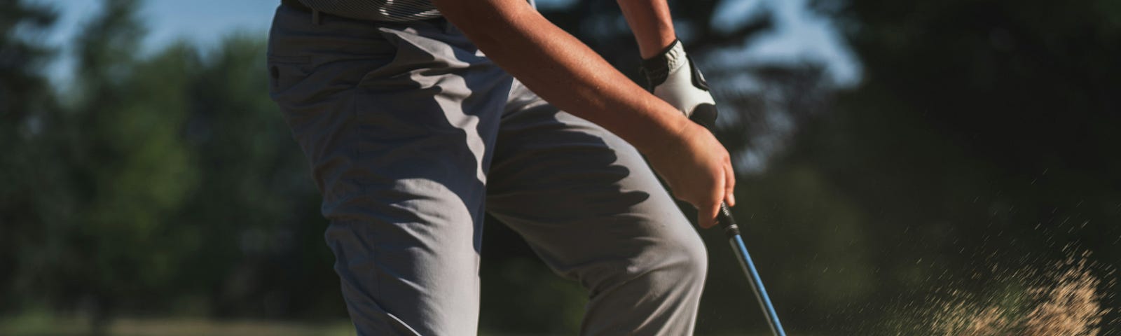 A golfer in gray golf pants whacks a golf by out of a sand area as sand flies in the air.