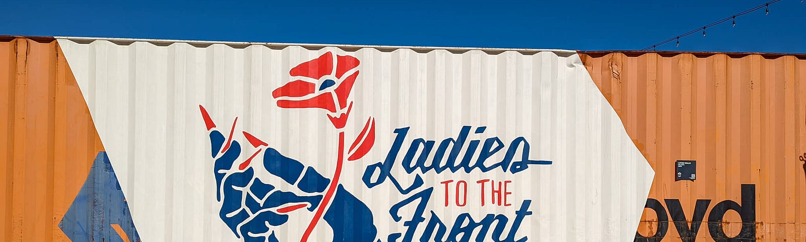 A shipping container painted with a large mural showing a hand holding a poppy flower and the slogan “Ladies to the Front”