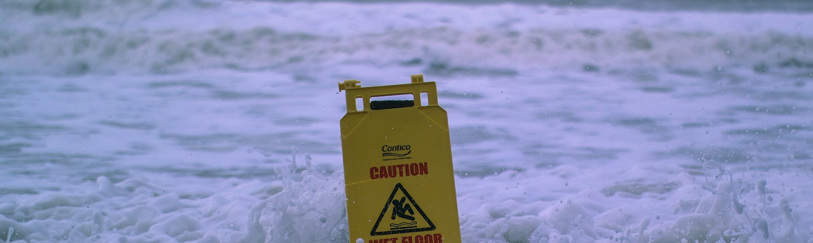 Yellow caution sign getting soaked by ocean on rocky beach.