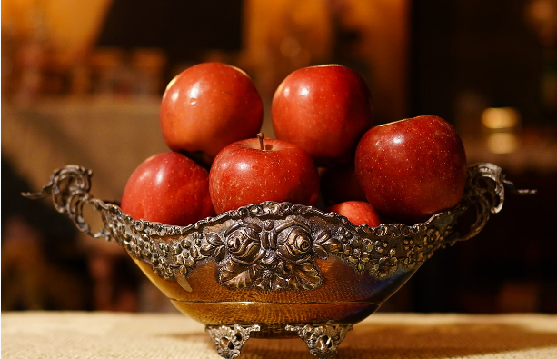 A photograph of red apples stacked in a footed, decorative silver bowl sitting on a light brown material. The background has been blurred.