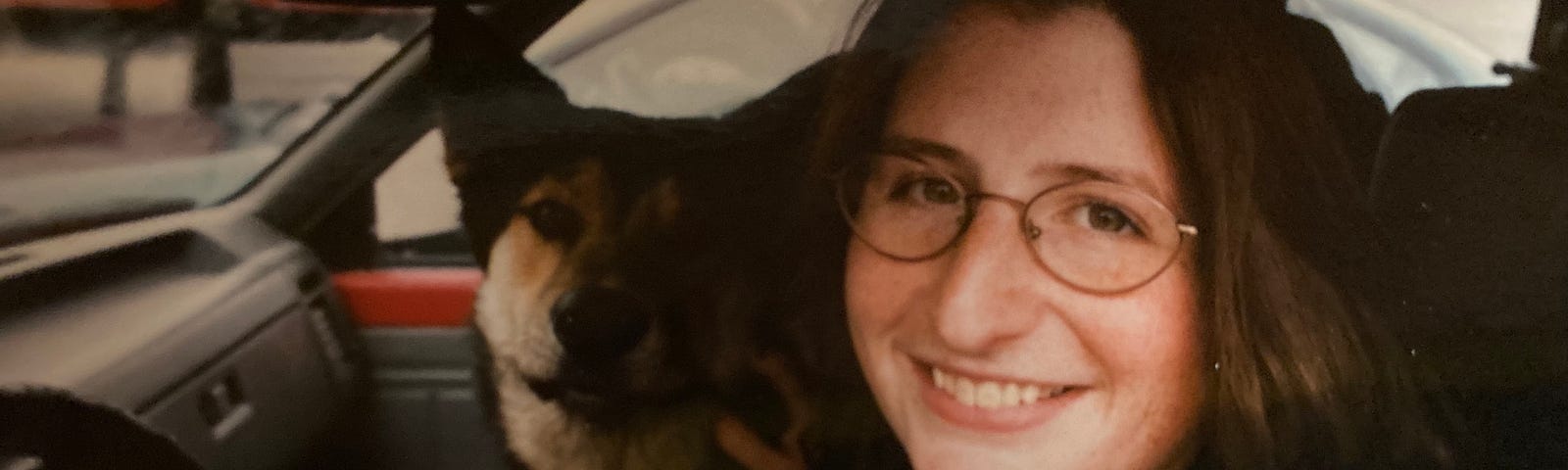 A young White woman with long brown hair and round glasses smiles at the camera from the driver’s seat of a truck. A German-shepherd husky mix dog is seated beside her in the passenger seat.