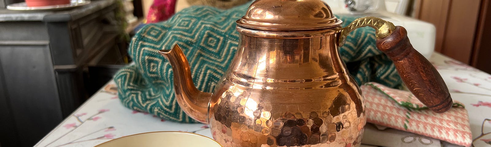 A vintage teacup full of tea sits on a sewing table next to a copper teapot