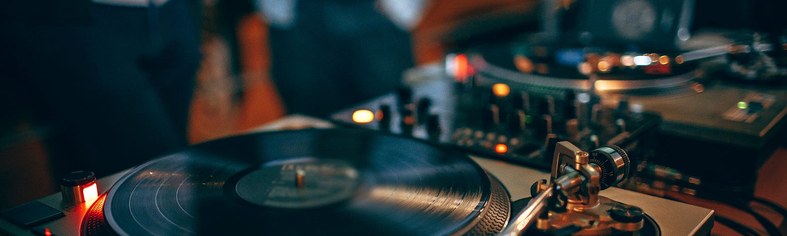 DJ station with 2 turntables in foreground and blurred figures congregating in background.