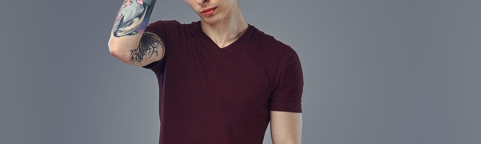 Handsome fashionable young guy with stylish hair and tattoo on his arm posing in a studio against a grey background.