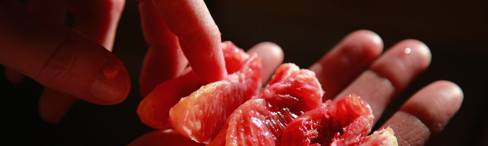 A carelessly sectioned blood-orange, ripped in places, held in one palm while the finger of the other hand prods it.