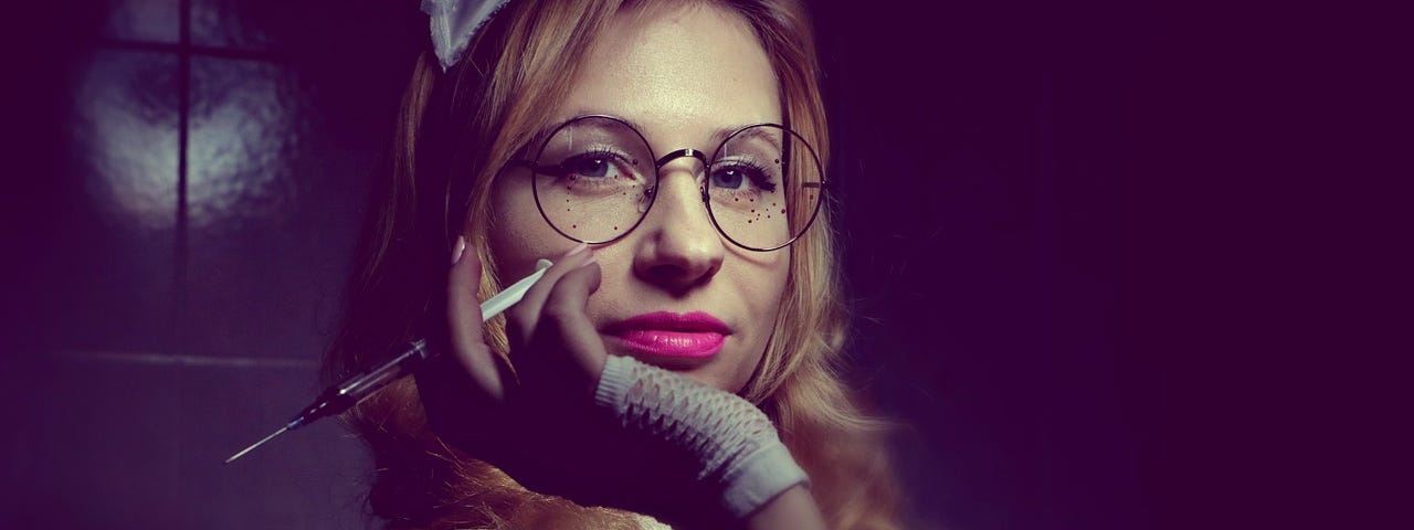 Image of a woman with an old-fashioned nurse’s cap on her head, resting her chin in her hand and in her hand she holds a syringe. She wears glasses and looks straight into the camera with a slight smile around her red lips.