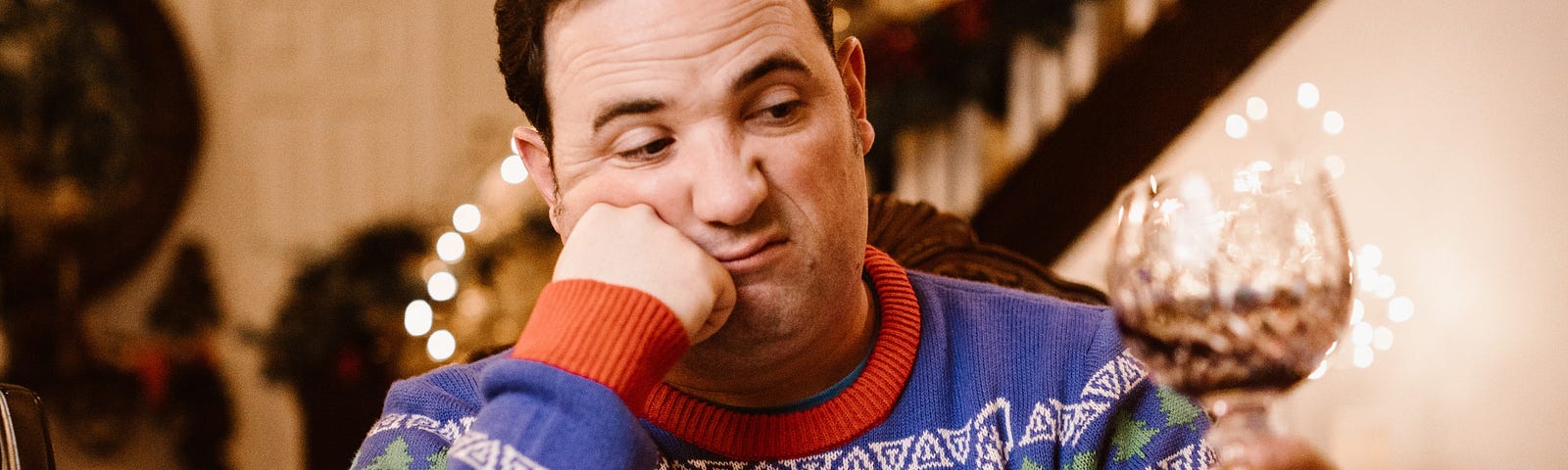 A man wearing a blue, white and red Christmas jumper. He has one fist against his cheek and looks grumpy.