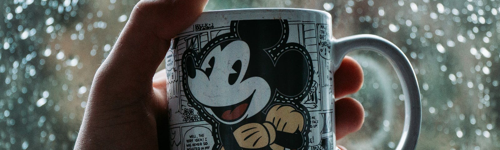 A hand holding a Disney Mickey Mouse mug in the foreground, with a rain-soaked windowpane in the background.