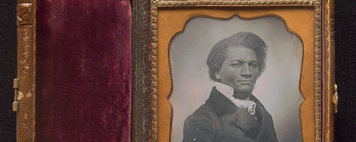 A black and white photographic portrait is displayed in a closable frame with a gold clasp. The frame is open. The right side of the frame is ornate in yellow and gold and contains a picture of a Black man wearing a formal black jacket with a formal white shirt underneath. He looks directly at the camera with a serious expression. The left side of the frame is red velvet.