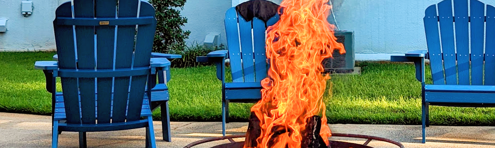 A fire pit with stars and moons on it roaring with fire and 3 blue adirondack chairs sit in the background on a patio close to a green patch of grass and white building