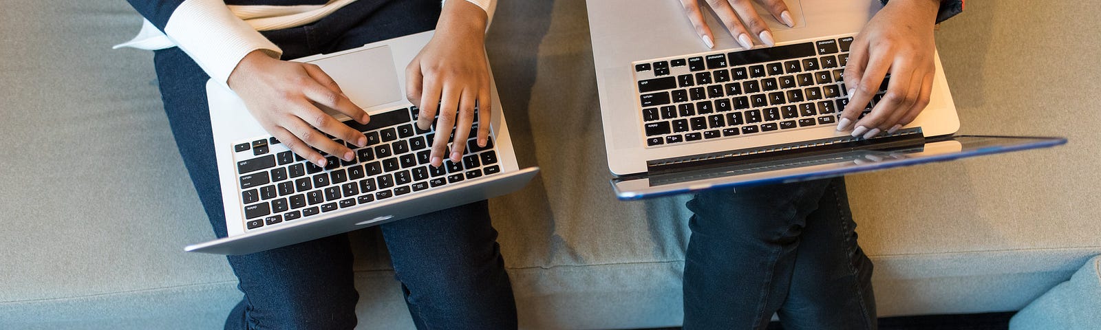 Photo from above of two women of color sitting on a couch, typing on macs.