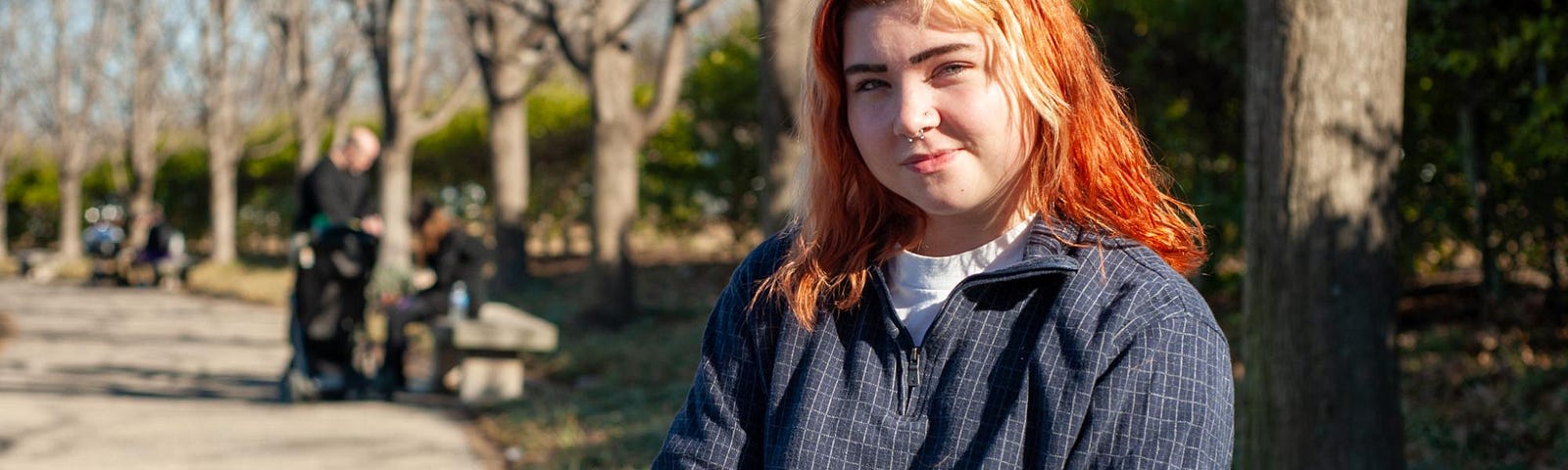 A smiling young white woman with orange and blonde hair and brown eyebrows. She is sitting on a bench in Forest Park with tree trunks in the background and is wearing a zip up long sleeved shirt that is navy with gray cross hatching pattern. She is smiling with a closed mouth and her eyes look kind.