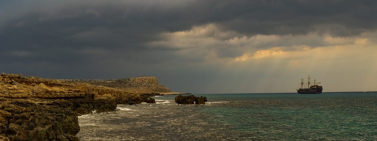 A rocky coast line and the ocean. An old wooden sailing ship is seen in the background. The skies are filled with dark rain clouds.