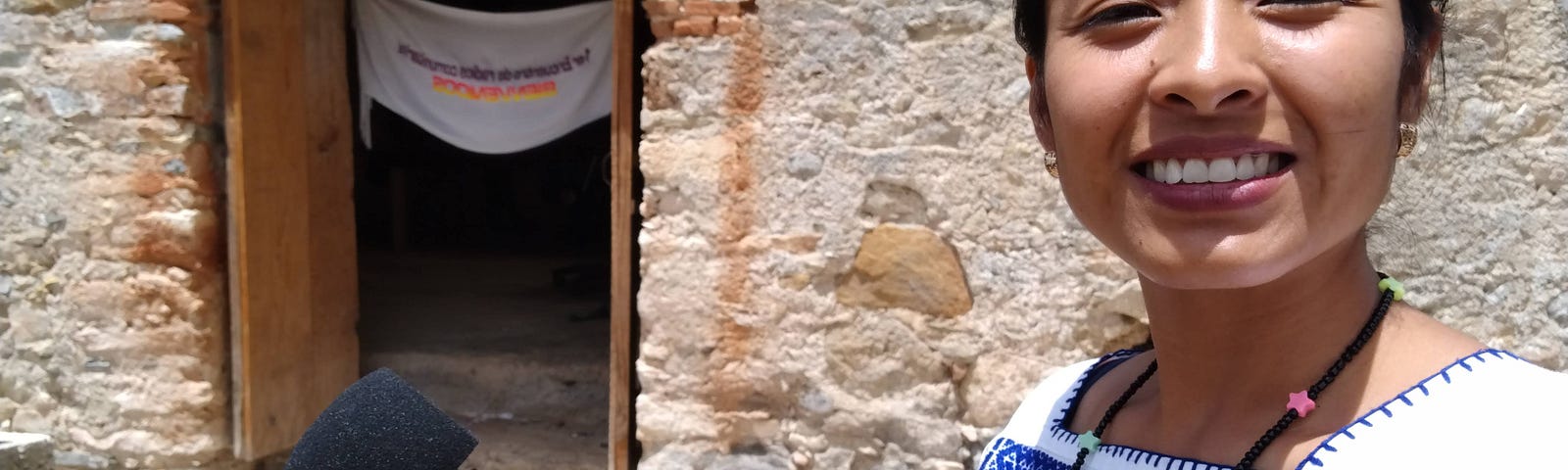 A woman journalist stands with a microphone outside a rural stone house while on a reporting trip in Oaxaca, Mexico.