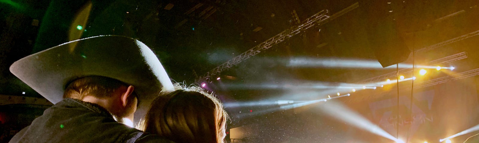 Two people standing in a crowd watch a concert. One’s wearing a cowboy hat.