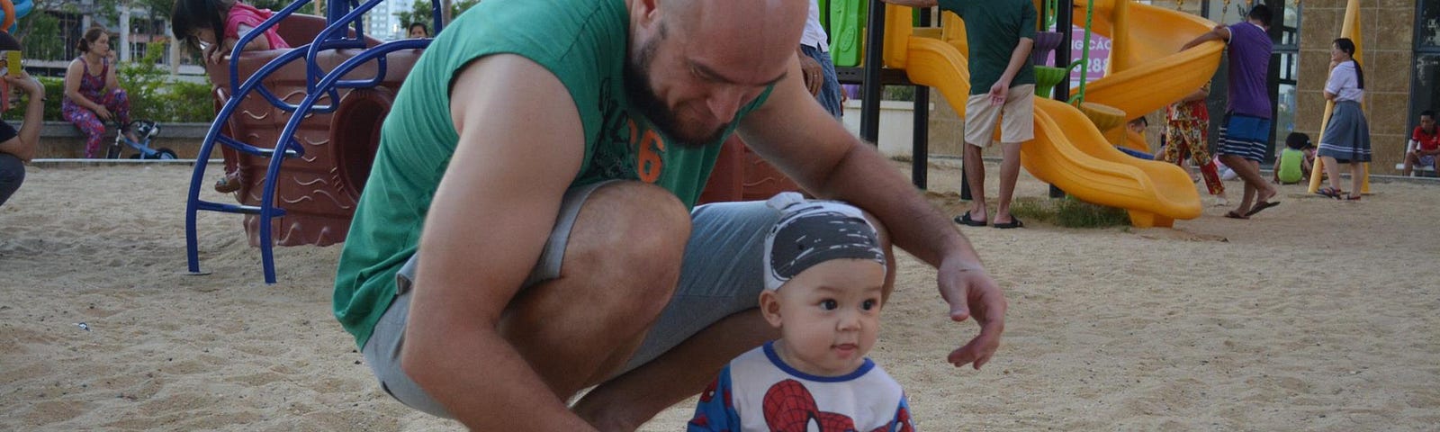 A man is squatting with a toddler boy in a sandbox