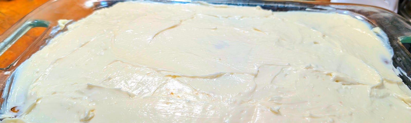 A glass baking dish is shown filled with a creamy white mixture, which appears to be either frosting or a dessert base not yet finished. The surface is uneven and suggests that the mixture has been spread manually with some kind of spatula.