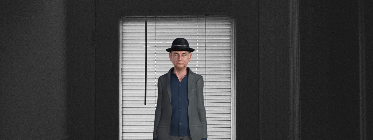 An old man in a grey suit, navy open-necked shirt, and black bowler hat standing in a darkened room in front of a backlit window with a venetian blind.