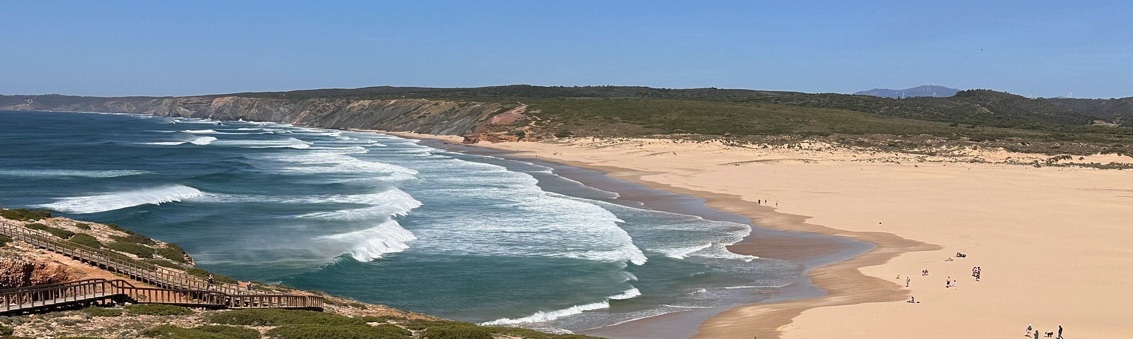 The beach in Borderia, Portugal. White waves sweeping into the sandy beach. Hills with spring flowers. People walking on the beach.