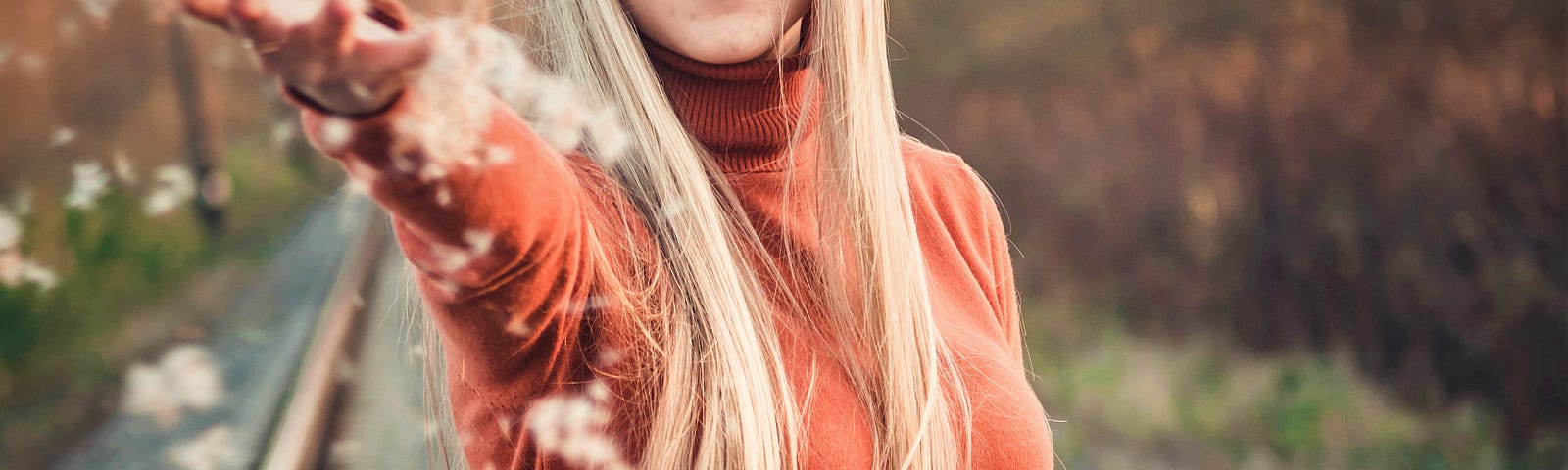 smiling girl with long blonde hair wearing long sleeve orange sweater standing on railroad tracks