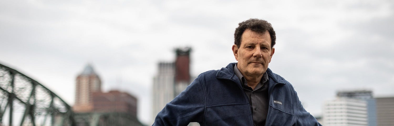 A man, Nicholas Kristof, standing on a bridge overpass, with downtown Portland, Oregon in the background.