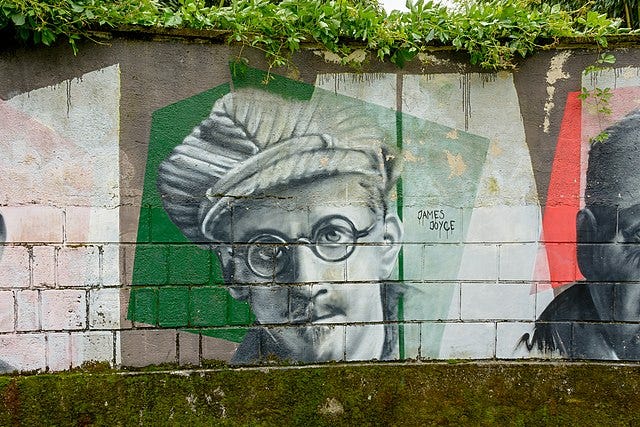 This image depicts a mural of a man with distinctive round glasses and a hat, portrayed in shades of gray with hints of green, against a concrete wall. The mural includes the name “JAMES JOYCE” alongside the portrait, indicating the depicted individual.