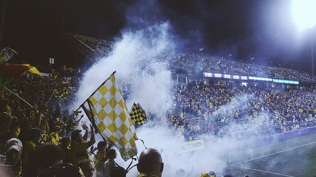 Soccer fans supporting their team during a soccer match. The atmosphere seems quite animated and wild, with smoke bombs, flags, and most likely sassy chants.