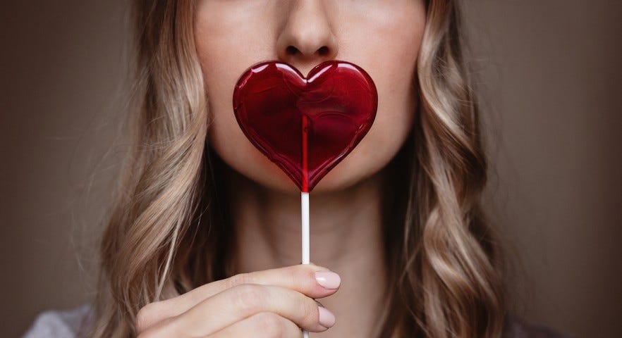 A woman with long hair holds a heart-shaped lollipop in front of her mouth.