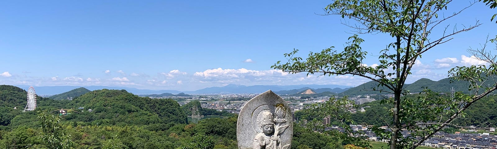 A layered view from a temple at the top of Mt. Tsugao. There is a bright blue sky with picturesque clouds and lush green mountains surrounding a small town in the background. In the foreground is the lookout spot from which the photo is taken, with square stone pillars and a statue of Bodhisattva sitting in front of a leaf-shaped wall with a snake carved into the block beneath him.