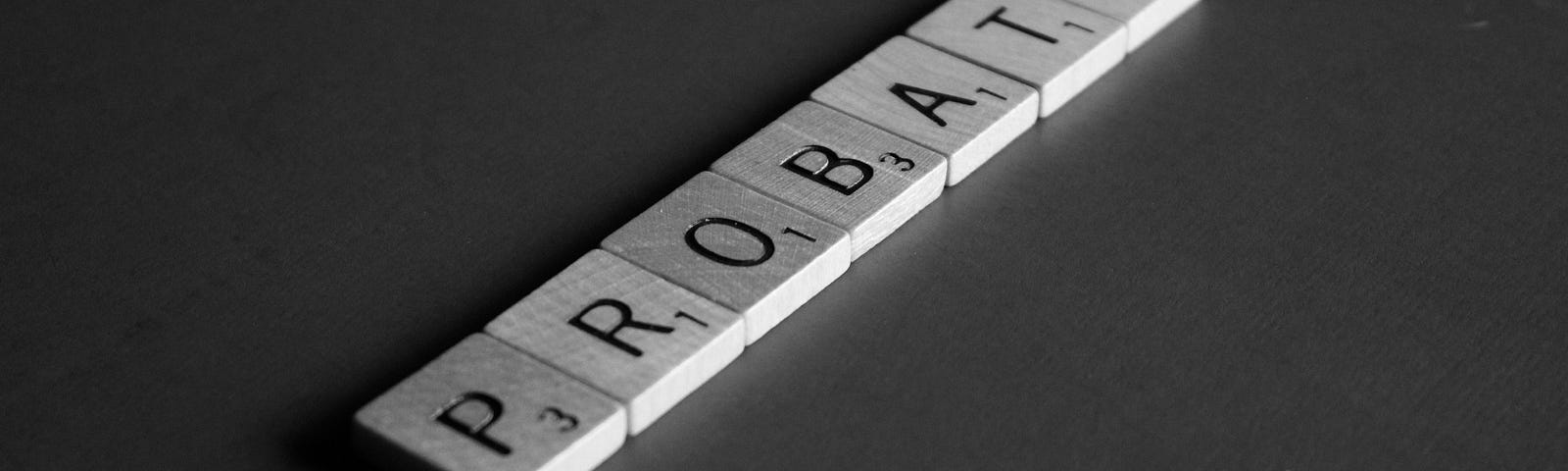 A black and white photo shows wooden Scrabble tiles that are placed on a flat surface. The letters on the tiles form the word “PROBATE.” The tiles are set at an angle.