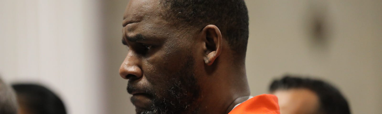 R. Kelly standing at a status hearing, wearing an orange prison jumpsuit