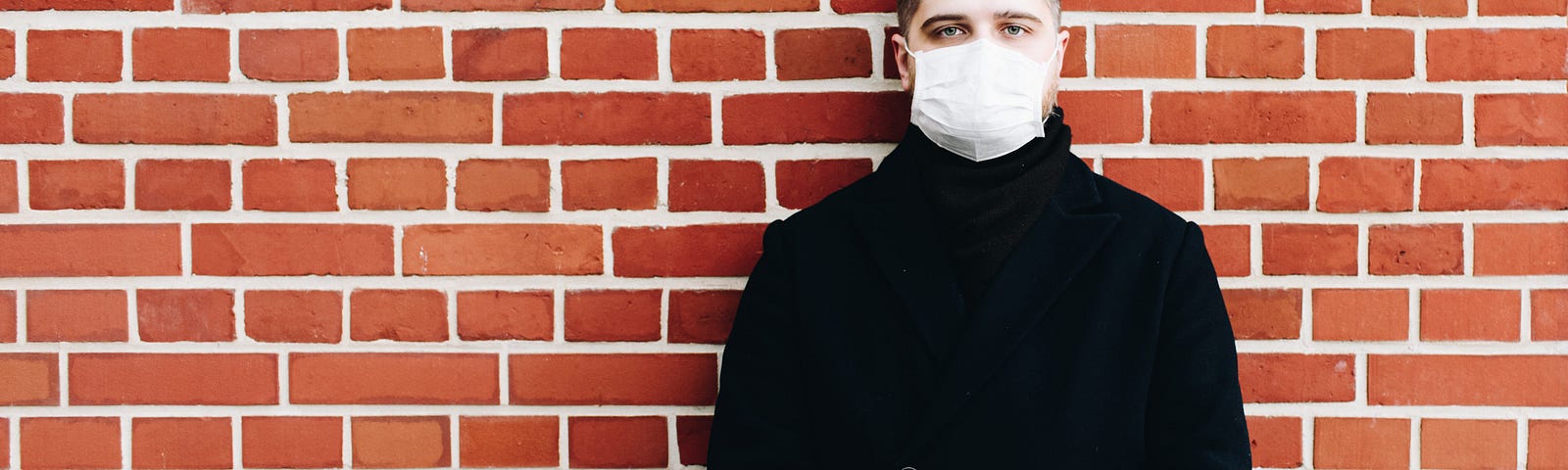 A man in a surgical mask stands against a brick wall.