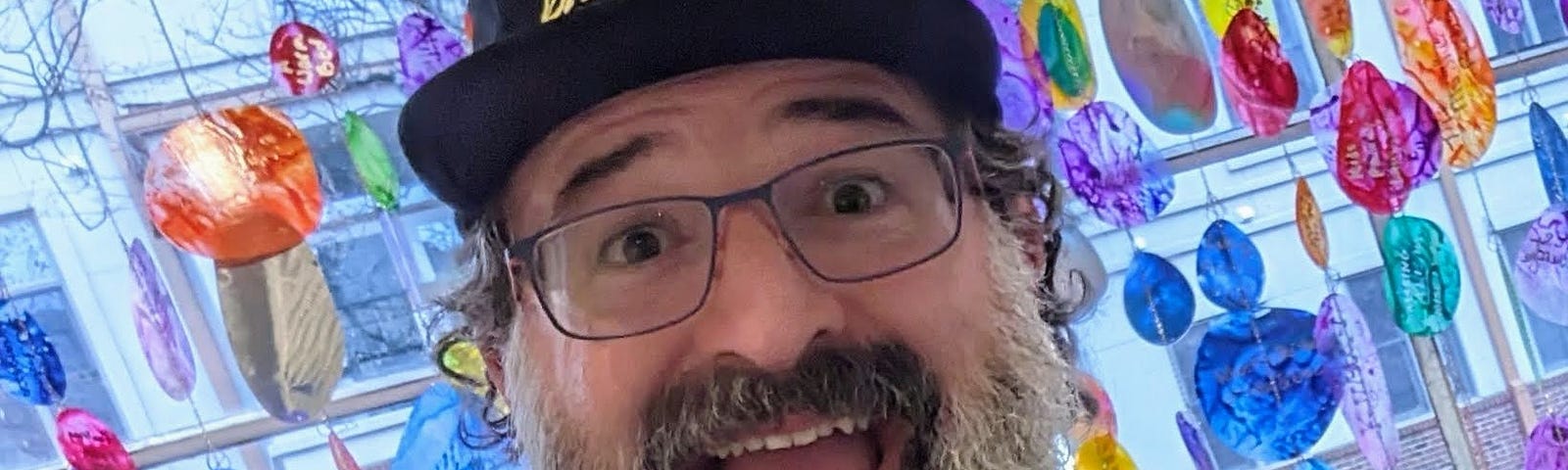 Closeup of the author with a big open-mouth smile. He’s wearing a hat, has a long graying beard and glasses, and is standing in front of colorful glass art.
